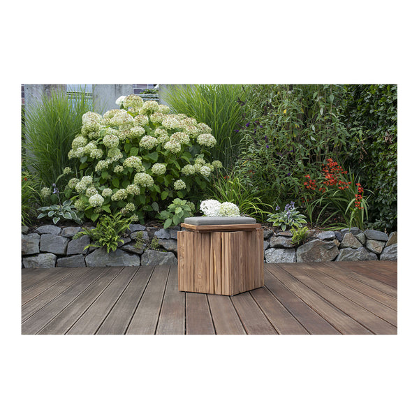 Ein Hocker Oxford-Holzhocker mit Kissen und Blumen darauf steht auf einem Deck, umgeben von üppigen Pflanzen und Felsen, und zeigt die Schönheit der Teak-Holz-Handwerkskunst.