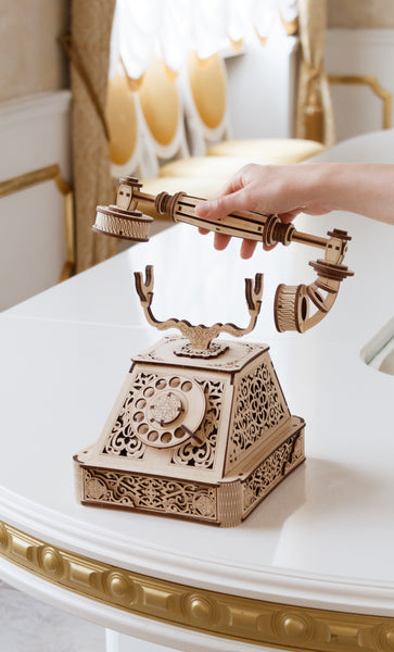 Eine Hand hebt den Hörer eines kunstvoll geschnitzten Retro-Telefons aus Holz von einem weißen Tisch ab.
