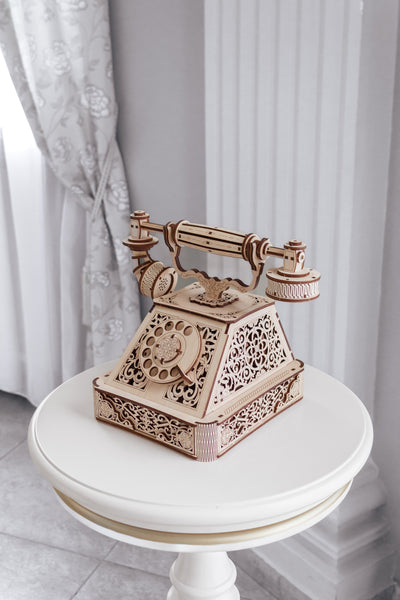 Ein kunstvoll detailliertes  antikes Telefonmodell aus Holz steht auf einem runden weißen Tisch vor einem eleganten Raumhintergrund mit Vorhängen und einer Säule.
