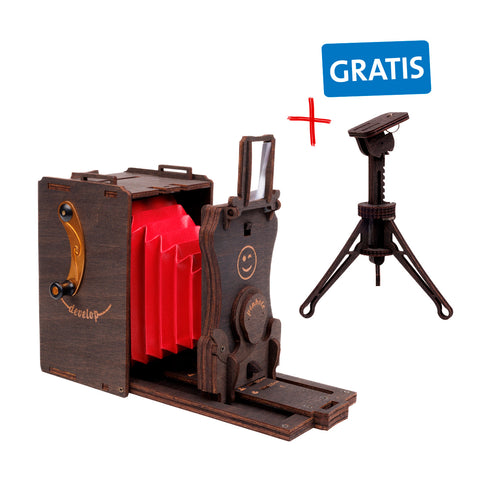 Eine nostalgisch aussehende Kamera aus Holz mit einem roten Balg, Gravuren und einem kleinen Drehhebel an der Seite. Daneben ein gemaltes Plus sowie ein Störer mit der Aufschrift Gratis und ein Stativ.
