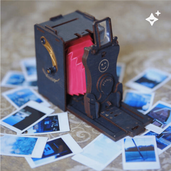 Ein dekoratives  miniaturhaftes  nostalgisches Kameramodell mit rot ausgezogener Balgenoptik steht auf einem Tisch  umgeben von verstreuten Polaroid-Fotos.
