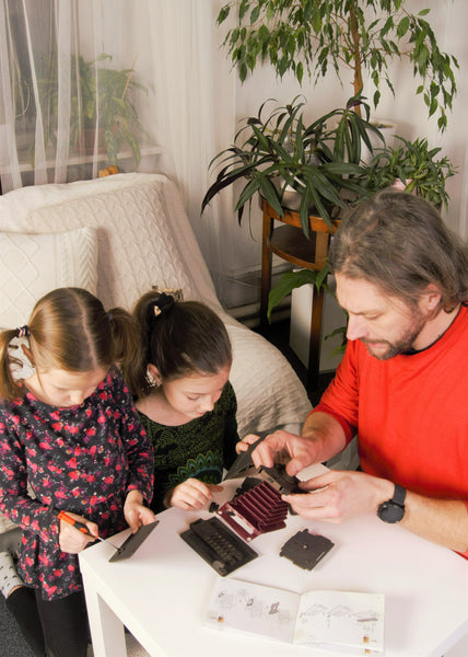 Ein Mann und zwei Kinder sitzen an einem weißen Tisch und arbeiten gemeinsam an einem Bastelprojekt  umgeben von Zimmerpflanzen in einem Wohnzimmer.
