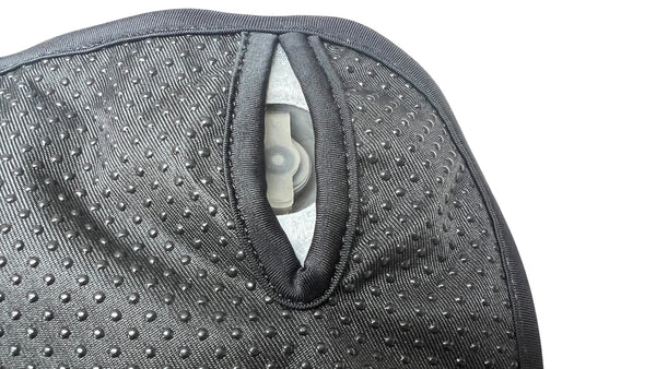 Nahaufnahme eines schwarzen  texturierten Materials mit einer ovalen Öffnung  die einen Teil eines dahinterliegenden mechanischen Teils zeigt.
