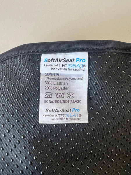 Ein schwarzer Stoff mit kleinen Löchern und einem angenähten weißen Etikett  auf dem Informationen über das Material des Produkts "SoftAirSeat Pro" von Tecseat stehen  darunter 50% TPU  30% Elasthan und 20% Polyester.
