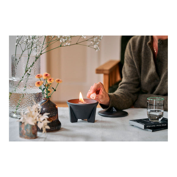 Eine Hand hält einen kleinen Wachswürfel über einen schwarzen Teelichthalter mit brennender Flamme  umgeben von rosa Blütenblättern auf einem Holztisch  im Hintergrund sind unscharfe bunte Kissen zu sehen.
