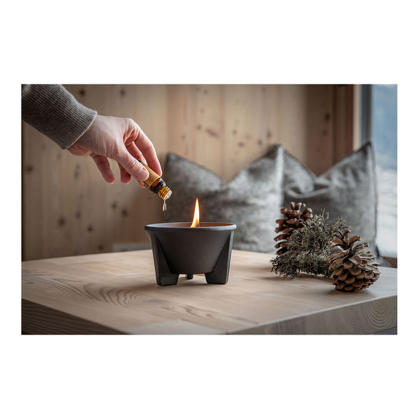 Eine Hand tropft Flüssigkeit aus einer kleinen Flasche in eine brennende Schale auf einem Holztisch  daneben liegen Tannenzapfen und Moos; im Hintergrund sind Kissen zu sehen.
