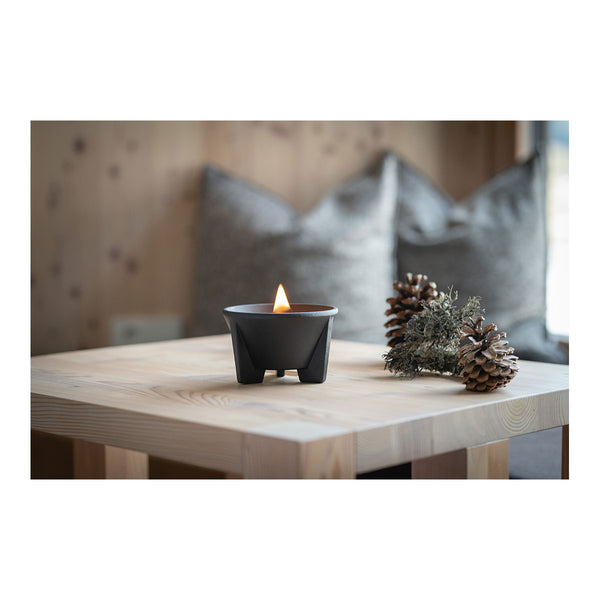 Ein kleines  schwarzes Teelicht brennt auf einem Holztisch neben Kiefernzapfen und Moos  mit verschwommenen  grauen Kissen im Hintergrund.
