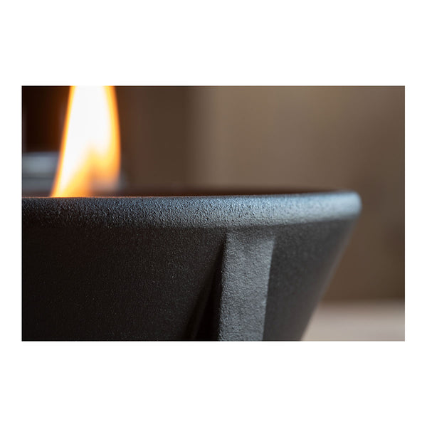 Eine Hand tropft Flüssigkeit aus einer kleinen Flasche in eine brennende Schale auf einem Holztisch  daneben liegen Tannenzapfen und Moos; im Hintergrund sind Kissen zu sehen.
