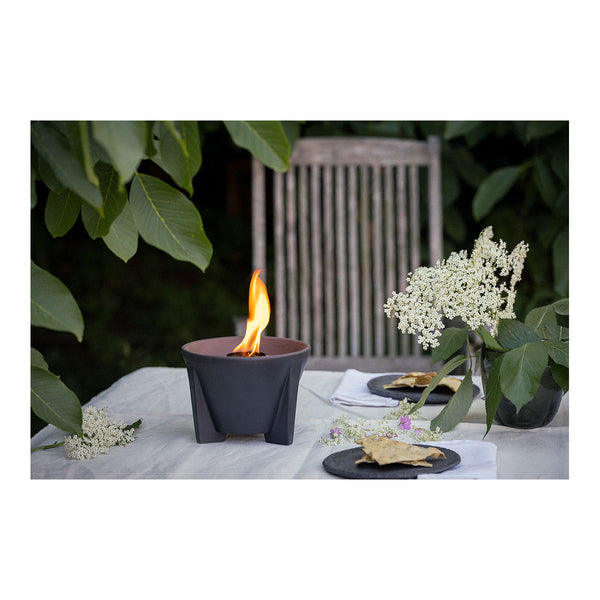 Ein Tisch im Freien mit einer brennenden Feuerstelle  einem Strauß weißer Blumen und tellern mit Knäckebrot  umgeben von üppigem Grün.
