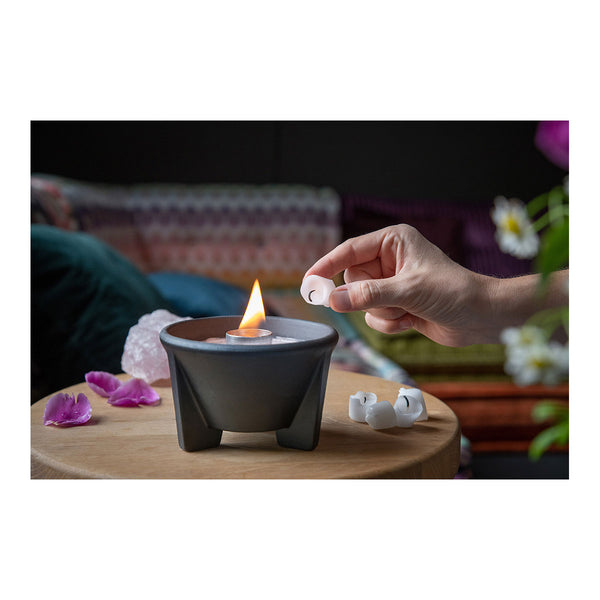 Eine Hand hält einen kleinen Wachswürfel über einen schwarzen Teelichthalter mit brennender Flamme  umgeben von rosa Blütenblättern auf einem Holztisch  im Hintergrund sind unscharfe bunte Kissen zu sehen.
