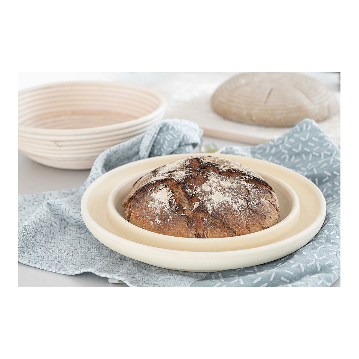 Ein gebackenes Rundbrot liegt auf einem Tuch in einer runden Holzform  mit einer weiteren leeren Gärform im Hintergrund.
