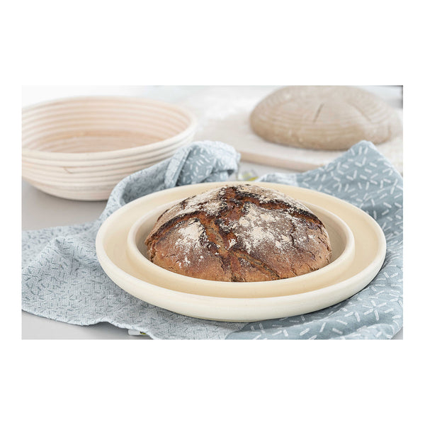 Ein gebackenes Rundbrot liegt auf einem Tuch in einer runden Holzform  mit einer weiteren leeren Gärform im Hintergrund.
