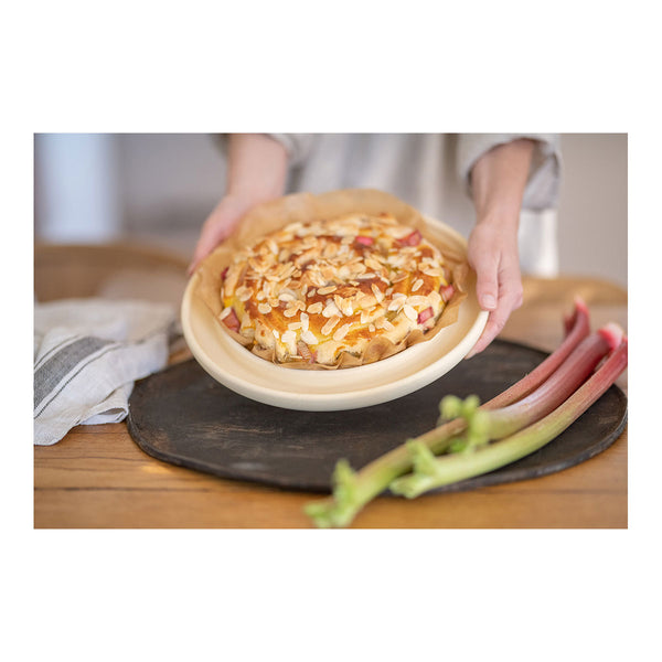 Eine Person hält einen frisch gebackenen Kuchen mit gehobelten Mandeln und Rhabarberstücken auf einem Tisch  daneben liegt frischer Rhabarber.
