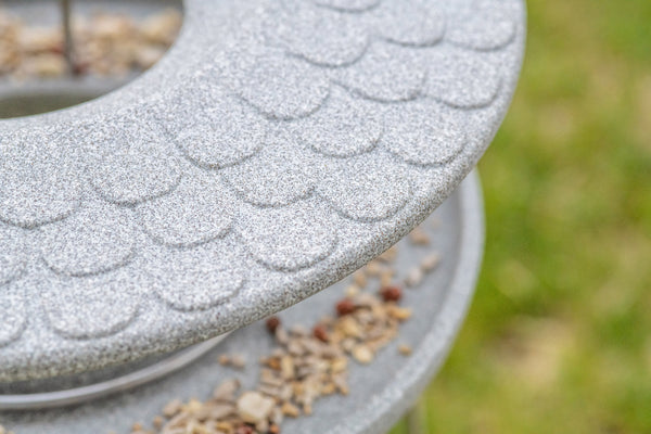 Nahaufnahme eines grauen Vogelhäuschens mit strukturierter Oberfläche  auf dem Vogelfutter verstreut ist  vor einem verschwommenen Gras-Hintergrund.
