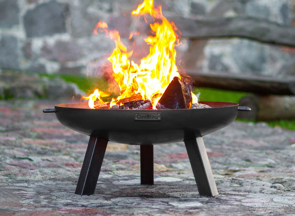 Eine schwarze Feuerschale steht auf einem gepflasterten Boden mit brennendem Feuer in der Mitte  umgeben von unscharfem Hintergrund mit Mauerwerk.
