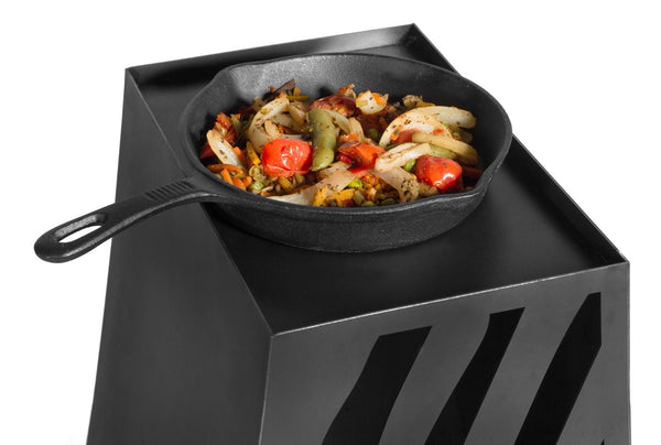 Eine gusseiserne Pfanne mit gebratenem Gemüse  darunter Zwiebeln und Paprika  steht auf einer schwarzen  würfelförmigen Oberfläche.
