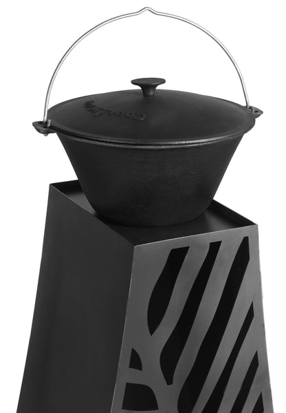 Ein schwarzer gusseiserner Topf mit Deckel und Tragegriff steht auf einem dekorativen schwarzen Sockel mit schrägen Streifenmustern.
