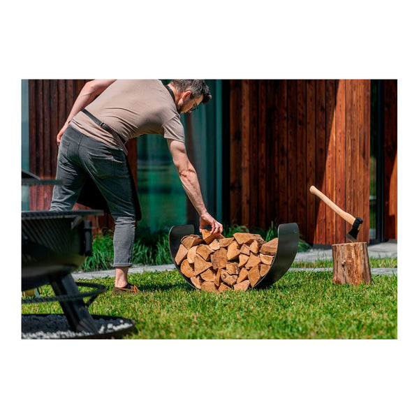 Ein Mann nimmt aus einem halbmondförmigen Regal ein Holzscheit raus. Daneben steckt eine Axt in einem Baumstumpf.