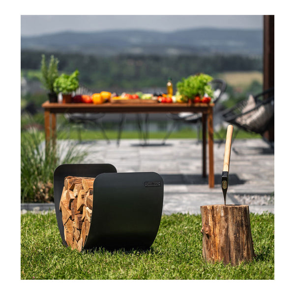 Ein schwarzer  halbkreisförmiger Holzkorb gefüllt mit gestapeltem Brennholz steht auf einem hellen Holzboden  im Hintergrund sind unscharf ein gedeckter Tisch mit Stuhl und Pflanzen zu sehen.
