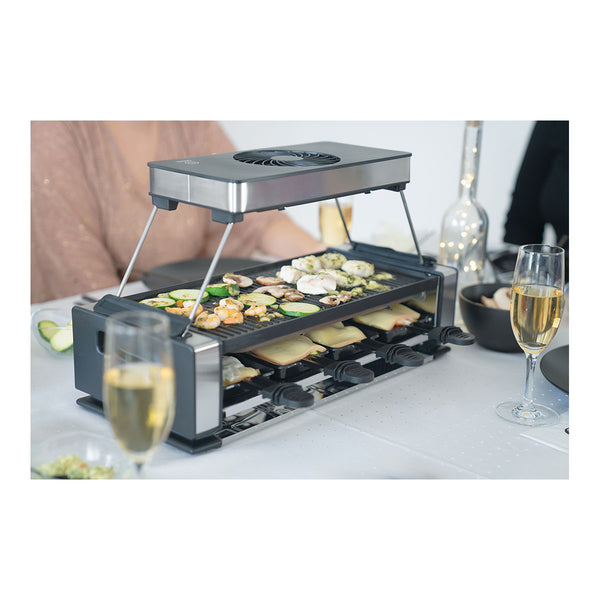 Ein eleganter Raclette-Grill auf einem gedeckten Tisch brät Gemüse und Meeresfrüchte auf der oberen Platte  während Käseschalen unten schmelzen; zwei Gläser Sekt stehen daneben.
