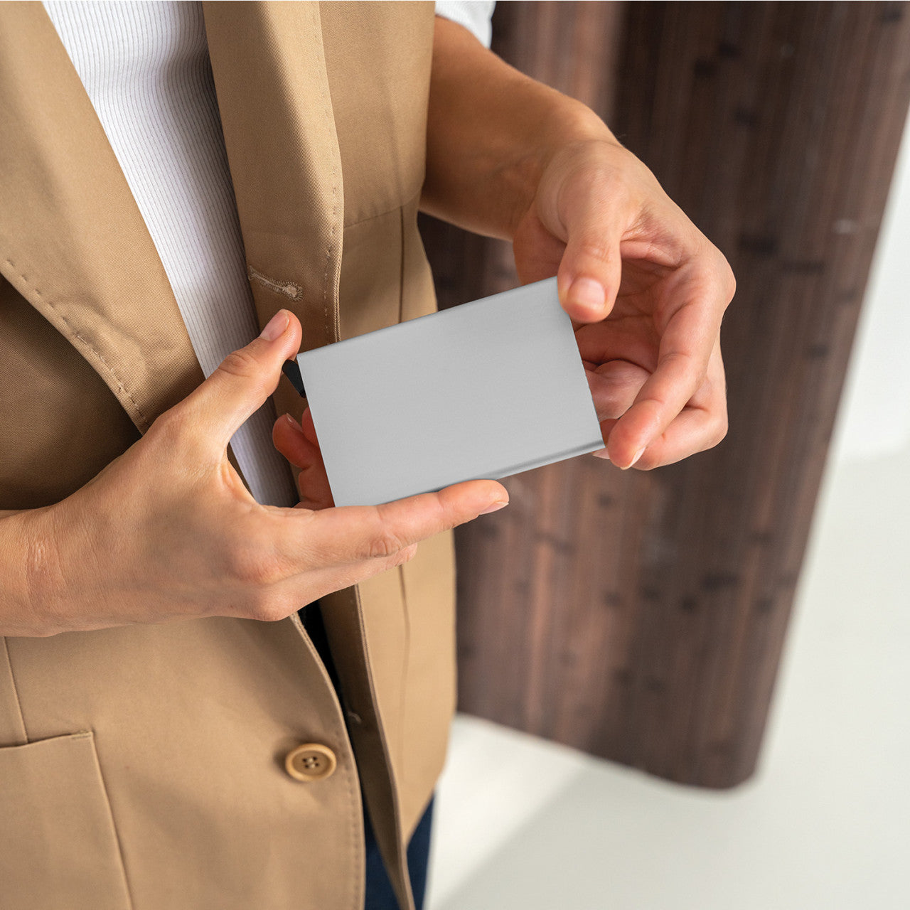 Person im hellbraunen Blazer, die vor einem hölzernen Hintergrund eine leere graue Visitenkarte in beiden Händen hält, perfekt für die Präsentation eines Aluminium-Kartenetuis mit RFID- oder NFC-Ausleseschutz.