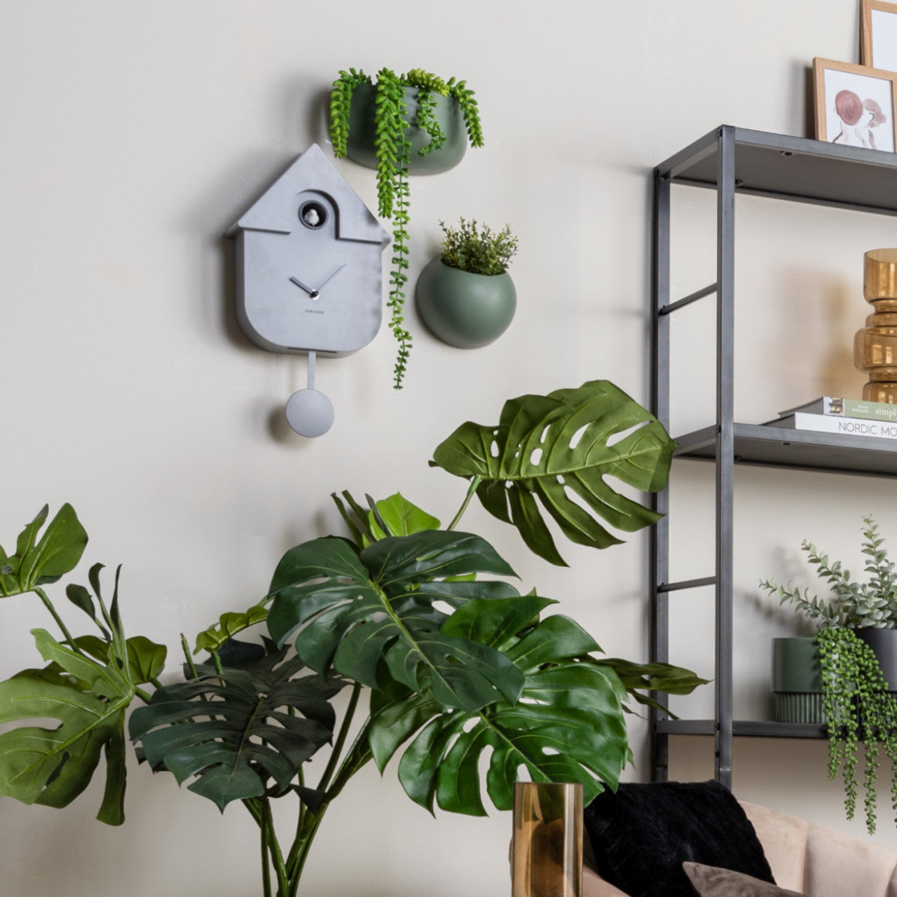 Modernes Wohnzimmer mit Grünpflanzen, einer grauen Kuckucksuhr in minimalistischem Design und einem Metallregal mit Dekor an einer beigen Wand.