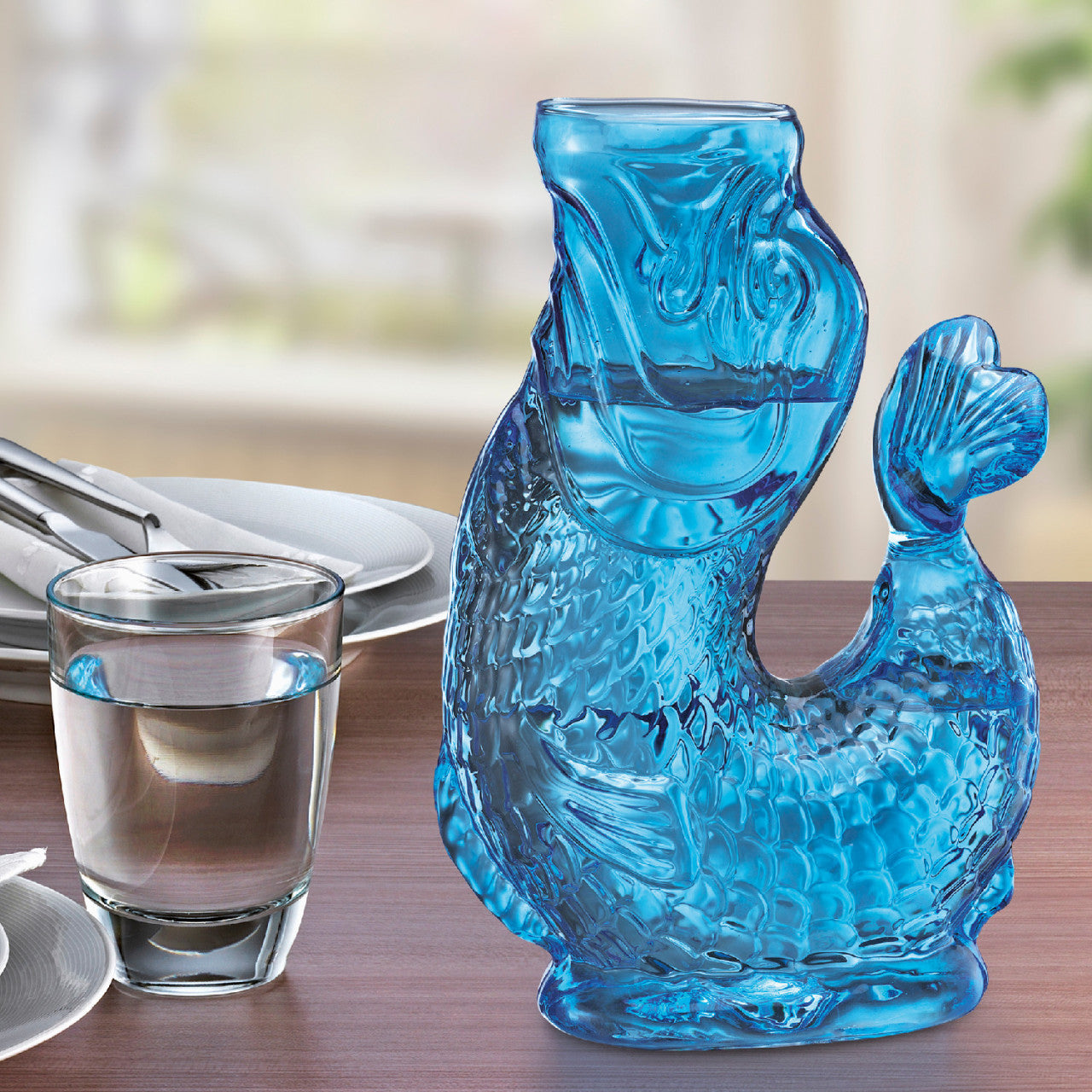 Eine blaue Fischform Wasserkaraffe aus Borosilikatglas steht auf dem Tisch neben einem Glas Wasser und Besteck.