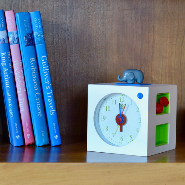 Ein Kinderwecker mit einem Elefanten oben drauf und Tierstimmen steht neben klassischen Büchern auf einem Holzregal.