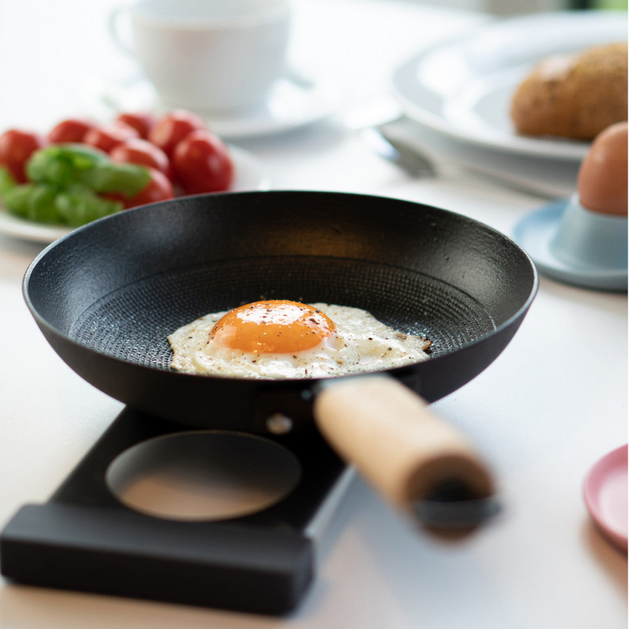 Ein Spiegelei wird in einer schwarzen Bratpfanne mit Edelstahleinsatz gebraten, im Hintergrund sind Frühstückszutaten unscharf.