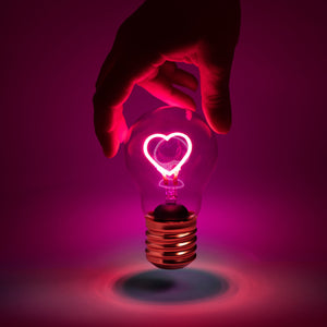 Eine Hand hält eine kabellose Glühbirne mit einem leuchtend rosa herzförmigen Glühfaden vor einem dunkelrosa Hintergrund.