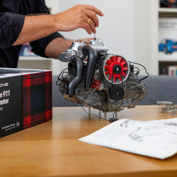 Eine Person baut auf einem Tisch einen maßstabsgetreuen Modellmotor zusammen, inspiriert von der Turbotechnologie von Hochleistungssportwagen wie dem Porsche 911 Turbo. In der Nähe befinden sich Anleitungen und eine Box.