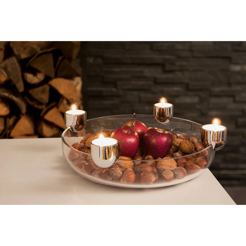 Eine Glasschale mit Äpfeln, Nüssen und vier brennenden Kerzen dient als moderner Adventskranz auf einem weißen Tisch vor einer dunklen Steinwand.