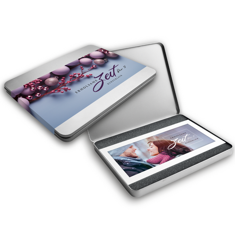 Geöffnete Metallbox in der ein Gutschein drin ist, auf dem ein Paar abgebildet ist. Daneben die Aufschrift "Erholsame Zeit". Daneben eine geschlossene Metallbox mit einem Deckel, auf dem ein lila-blaues Design mit Weihnachtskugeln und Beeren sowie dem Text "Erholsame Zeit für 2 – Kurzurlaub" abgebildet ist