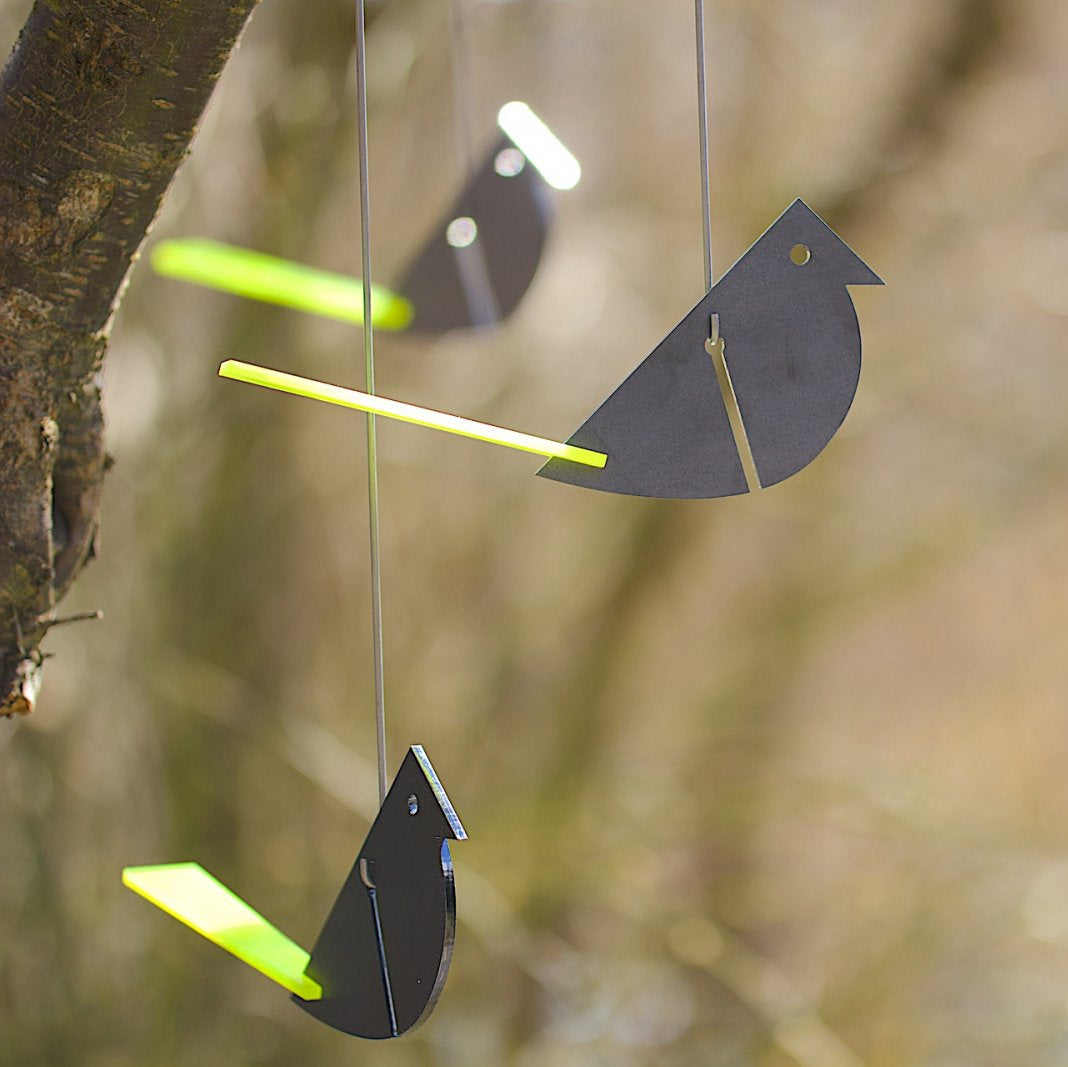 Papiervogel-Ornamente mit gelben Flügeln hängen an Schnüren an einem Ast im Freien und ähneln einem bezaubernden Gartenvogel-Windspiel. Der unscharfe Hintergrund hebt ihre zarten Bewegungen im Wind hervor.