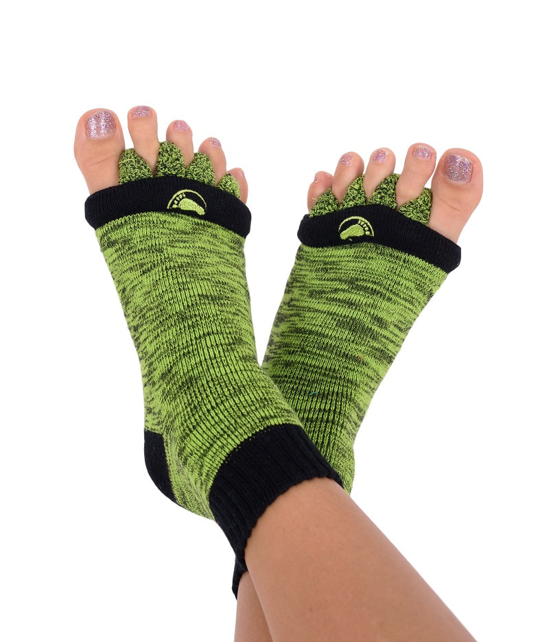 In grünen Fußausrichtungssocken mit schwarzem Besatz und glitzernden Zehennägeln lümmelt man herum, stellt das offene Zehendesign und das dezente Logo oben zur Schau und demonstriert den Komfort, den diese Socken bieten, wenn die Füße sanft an den Knöcheln überkreuzt ruhen.