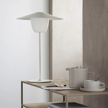 Minimalistischer Tisch mit mobiler LED-Leuchte mit integriertem Akku, moderner weißer Lampe, Teekanne, Tasse und gestapelten Büchern vor einer neutralen Wand – ideal für In-/Outdoornutzung.