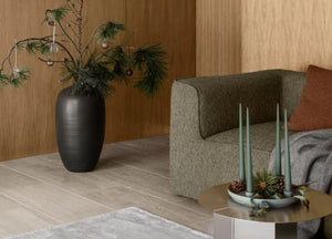 Moderne Wohnzimmerecke mit grünem Sofa, schwarzer Vase mit Tannenzweigen und Kerzen auf einem runden Tisch. Festlicher Weihnachtsbaumschmuck und eine schimmernde Glaskugel verleihen dem stilvollen Ambiente dezenten Weihnachtscharme.