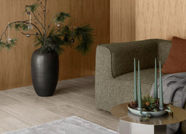 Moderne Wohnzimmerecke mit grünem Sofa, schwarzer Vase mit Tannenzweigen und Kerzen auf einem runden Tisch. Festlicher Weihnachtsbaumschmuck und eine schimmernde Glaskugel verleihen dem stilvollen Ambiente dezenten Weihnachtscharme.