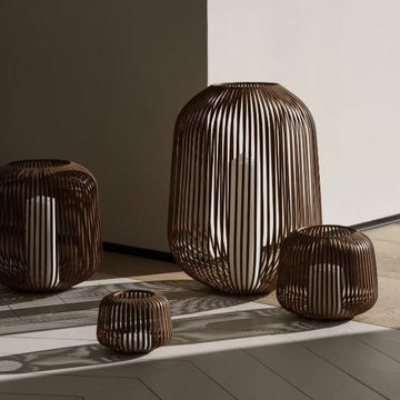 Vier braune LITO-Laternen aus Korbgeflecht mit weißen Kerzen darin, die Schatten auf einen hellen Boden und eine helle Wand werfen – perfekt, um in Ihrem Außenbereich eine gemütliche Atmosphäre zu schaffen.