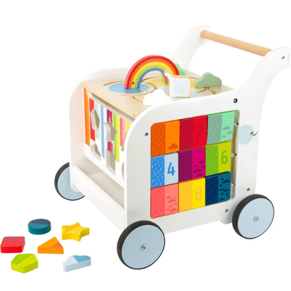 Weißer Lauflernwagen aus Holz mit bunten Bauklötzen, Formen, Motorikspielzeug und einem Regenbogenspielzeug auf vier Rädern – inklusive Tafelfläche für kreatives Spielen.