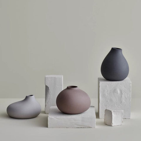 Drei minimalistische Keramikvasen, darunter die Vase NONA aus unglasiertem Porzellan, stehen auf weißen Sockeln vor neutralem Hintergrund und strahlen in schlichter Präsentation natürliche Eleganz aus.