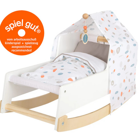 Weiße Puppenwiege aus Holz mit Stoffhimmel und Bettzeug, versehen mit einem Spiel Gut-Auszeichnungsaufkleber in der oberen linken Ecke – eine bezaubernde Wiege im skandinavischen Design für fantasievolles Spielen.