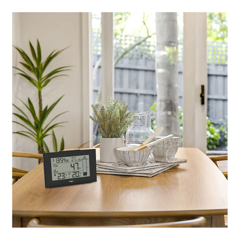 Eine Digitaluhr auf einem hölzernen Esstisch mit Schüsseln, einem Krug und einer Topfpflanze am Fenster, daneben ein Funk-Regenmesser zur kabellosen Übertragung der aktuellen Niederschlagsmengen.