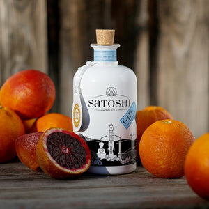 Eine weiße Flasche Satoshi Gin, hergestellt aus sizilianischer Blutorange und toskanischem Wacholder, steht zwischen ganzen und geschnittenen Blutorangen auf einer Holzoberfläche.