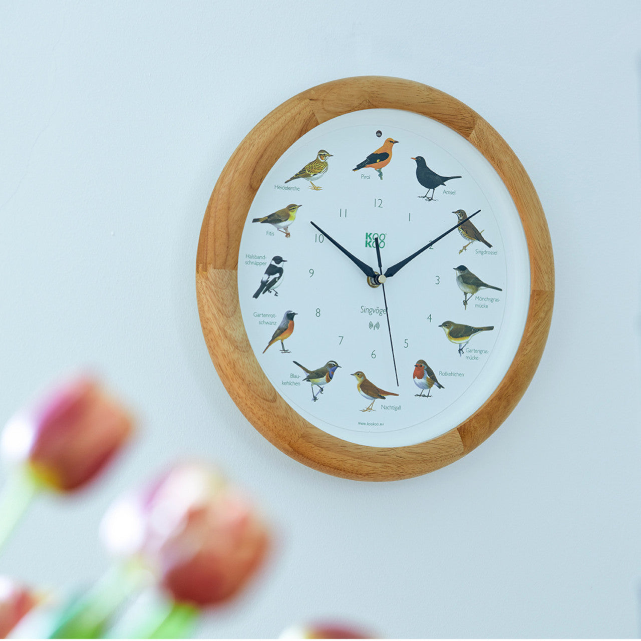 Runde Wanduhr mit Singvögel-Illustrationen für jede Stunde, Holzrahmen und verschwommenen Blumen im Vordergrund. Ausgestattet mit einem Lichtsensor, wie er auch in der KooKoo Vogelstimmenuhr-Serie zu finden ist.
