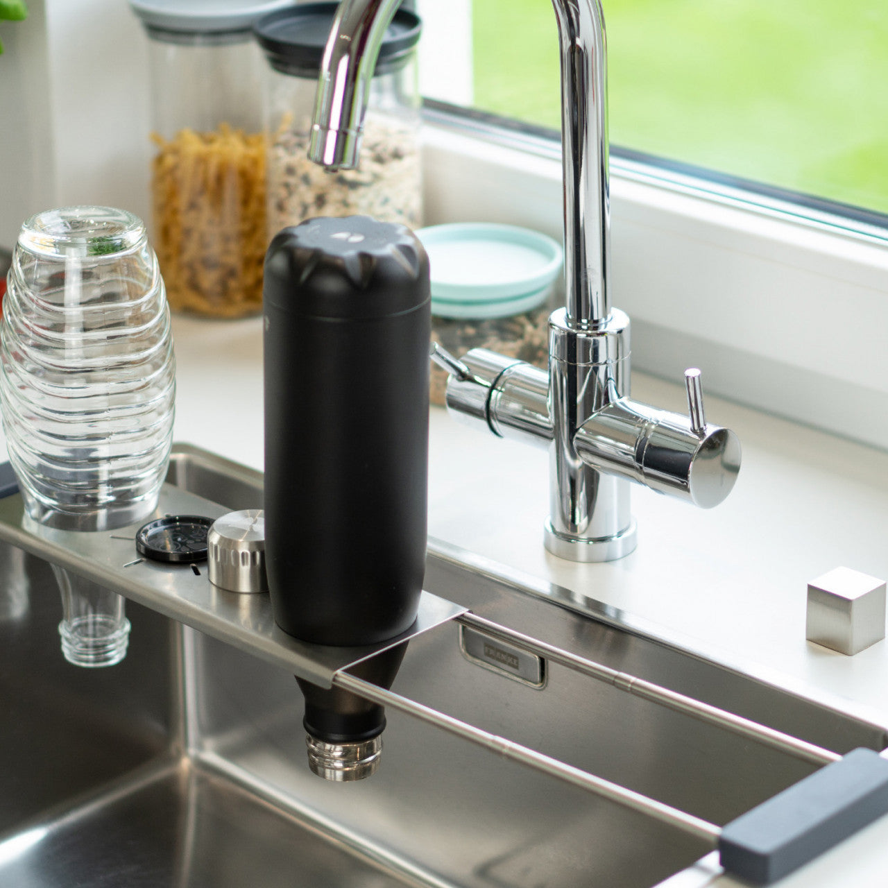 Eine schwarze Flasche trocknet kopfüber auf einer Drip.it-Abtropfstation neben einer Küchenspüle mit einem Chromhahn, Edelstahleinsatz und Glaswaren in der Nähe.