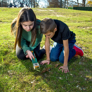 An einem sonnigen Tag knien zwei Kinder im Gras und untersuchen den Boden genau mit einer Acrylglas-Lupe.