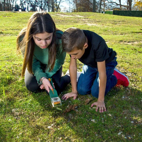 An einem sonnigen Tag knien zwei Kinder im Gras und untersuchen den Boden genau mit einer Acrylglas-Lupe.