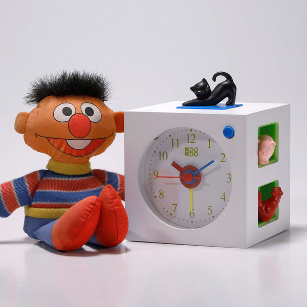 Die Plüschpuppe Ernie sitzt neben einem weißen Kinderwecker-Uhrenwürfel mit Tierfiguren, Tierstimmen und einer schwarzen Katze oben drauf.