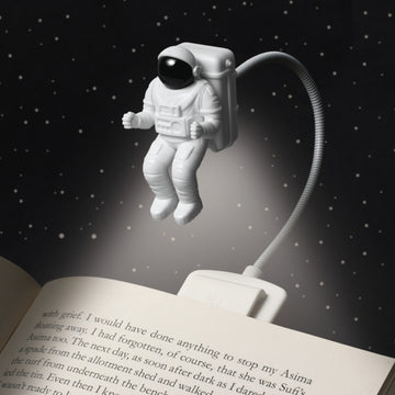 Eine kleine Astronauten-Leseleuchte schwebt über einem offenen Buch und ihr LED-Lichtstrahl beleuchtet die Seiten vor einem Sternenhintergrund.